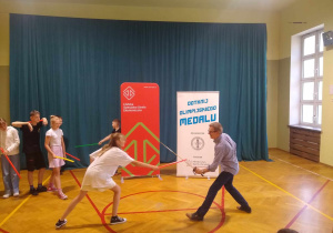 Uczennica w sali gimnastycznej walczy z olimpijczykiem na laskę gimnastyczną.