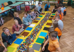 Uczniowie siedzą na podłodze przy rozłożonej żółtej macie, na której znajdują się klocki Lego.