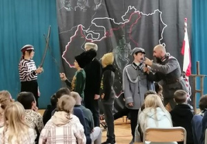 Zdjęcie przedstawia prowadzącego program Pana Michała, który rozmawia z uczniami biorącymi udział w inscenizacji.