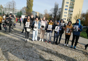 Zdjęcie przedstawia grupę ośmiu uczniów stojących przed Pomnikiem Pękniętego Serca. Każdy z nich trzyma w dłoniach zdjęcie dziecka, które w czasie II wojny światowej było więzione w Obozie koncentracyjnym dla dzieci przy ul. Przemysłowej.