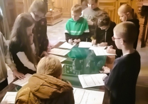 Zdjęcie przedstawia uczniów, którzy stojąc przy stole uzupełniają krzyżówkę.