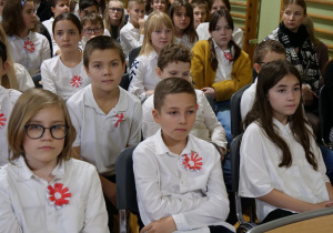 Uczniowie w strojach galowych siedzą w sali gimnastycznej i oglądają uroczystość z okazji Narodowego Święta Niepodległości.