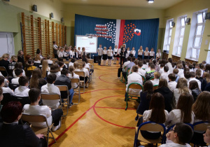 Uczniowie w strojach galowych siedzą w sali gimnastycznej i oglądają uroczystość z okazji Narodowego Święta Niepodległości.