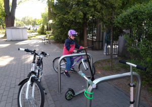 Na zdjęciu dziewczynka w kasku parkuje rower przy stojaku rowerowym.